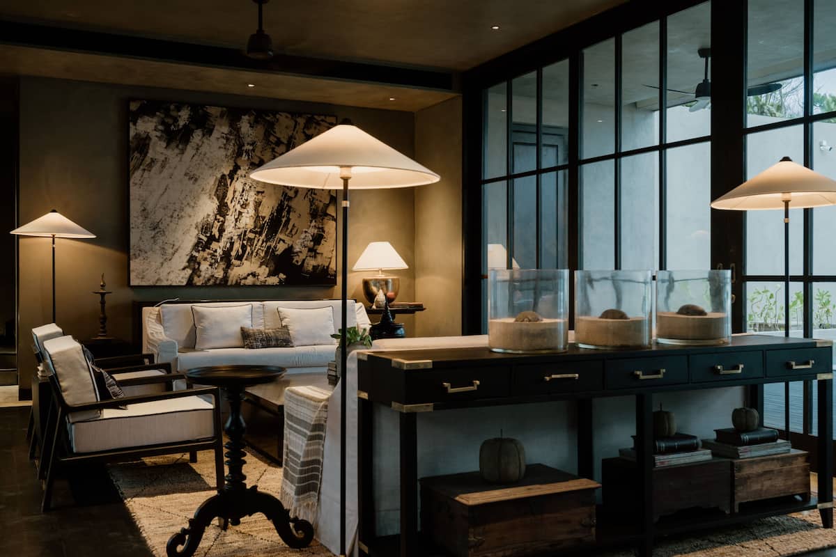 A modern living room with a sofa, armchairs, a coffee table, and a large window with a view. There are lamps on either side of the sofa and a decorative piece on the coffee table.
