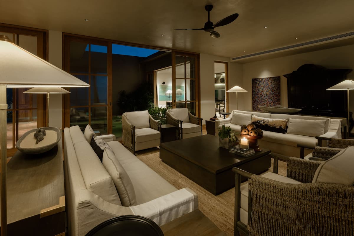 A modern living room with a sofa, armchairs, a coffee table, and a TV. There are lamps, a ceiling fan, and a view of the outdoors through large windows.
