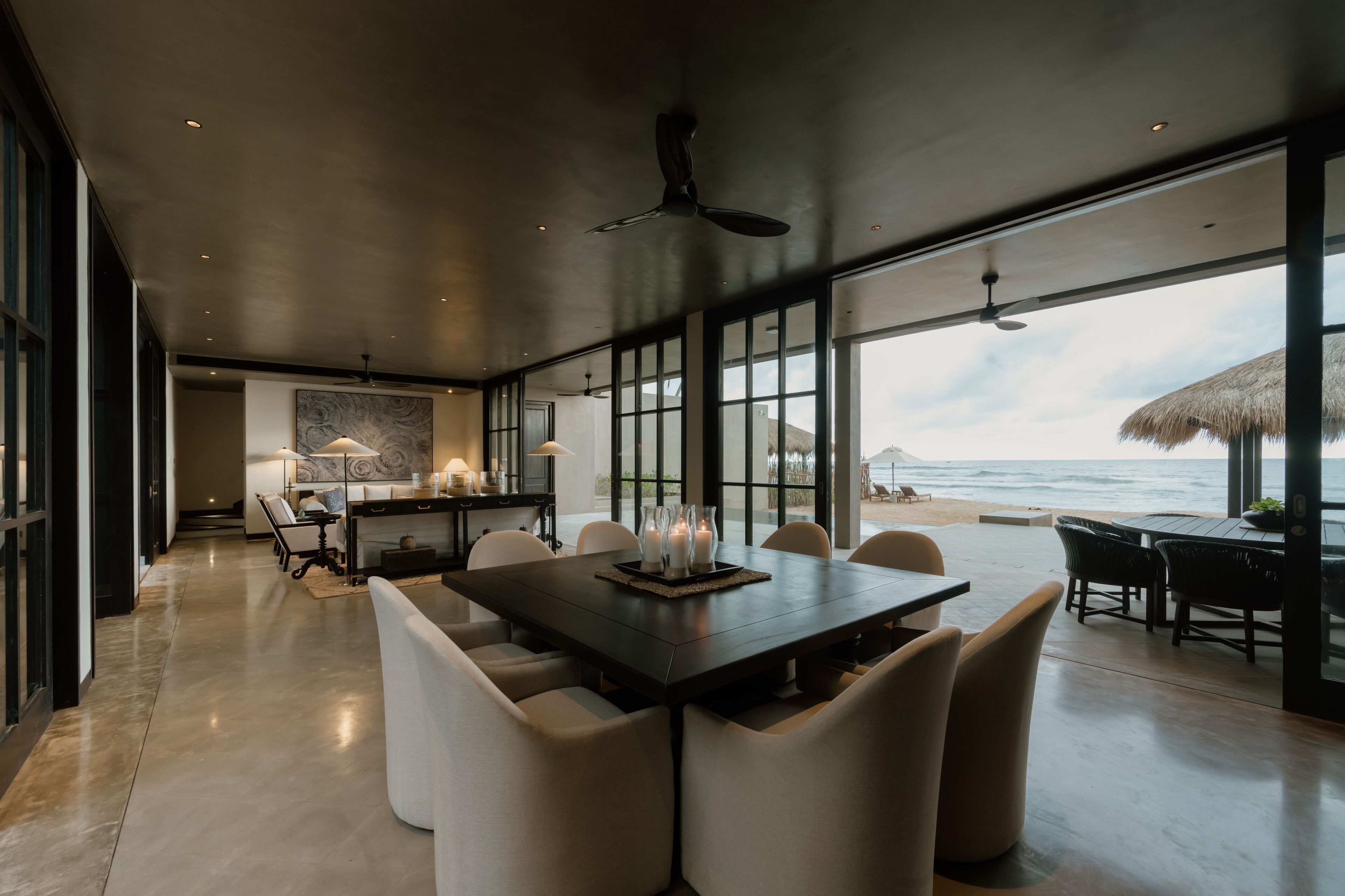 Un comedor moderno con una gran mesa y sillas, ventanas amplias con vistas al océano y un ventilador de techo.