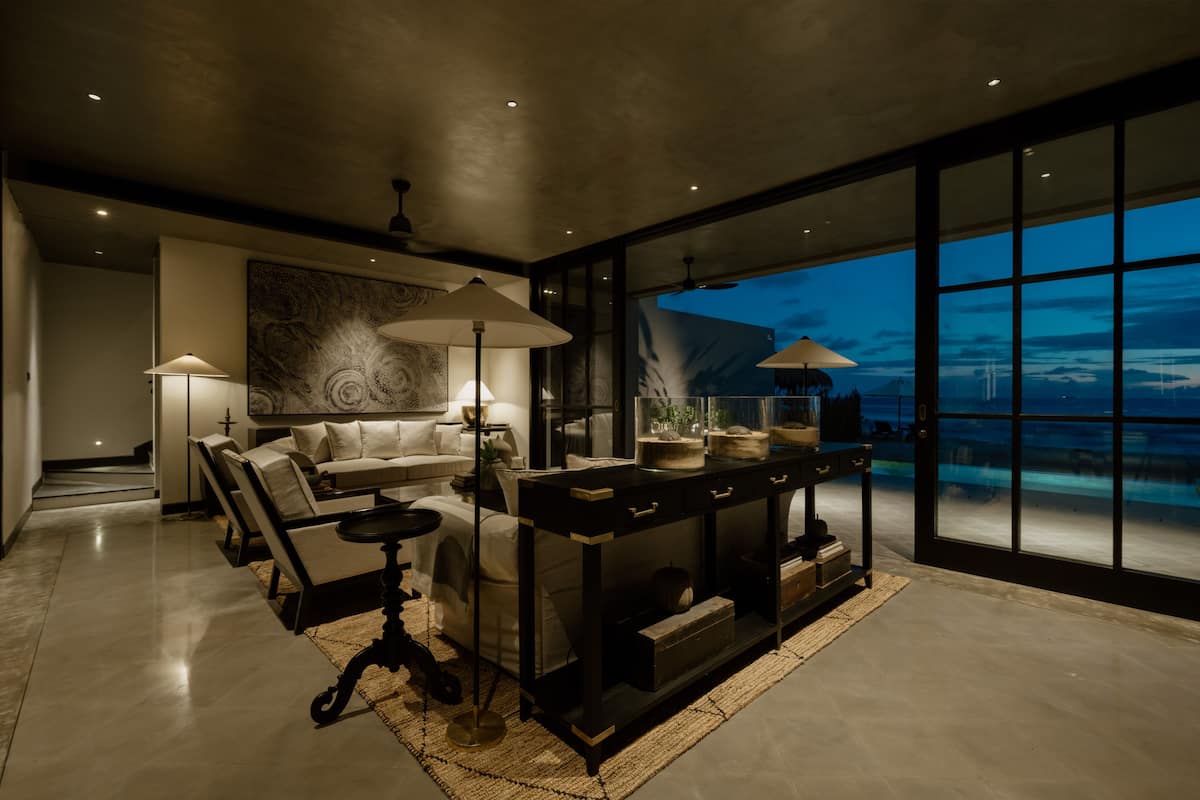 A modern living room with a sofa, coffee table, and lamps. There is a large window with a view of the sky and a pool.