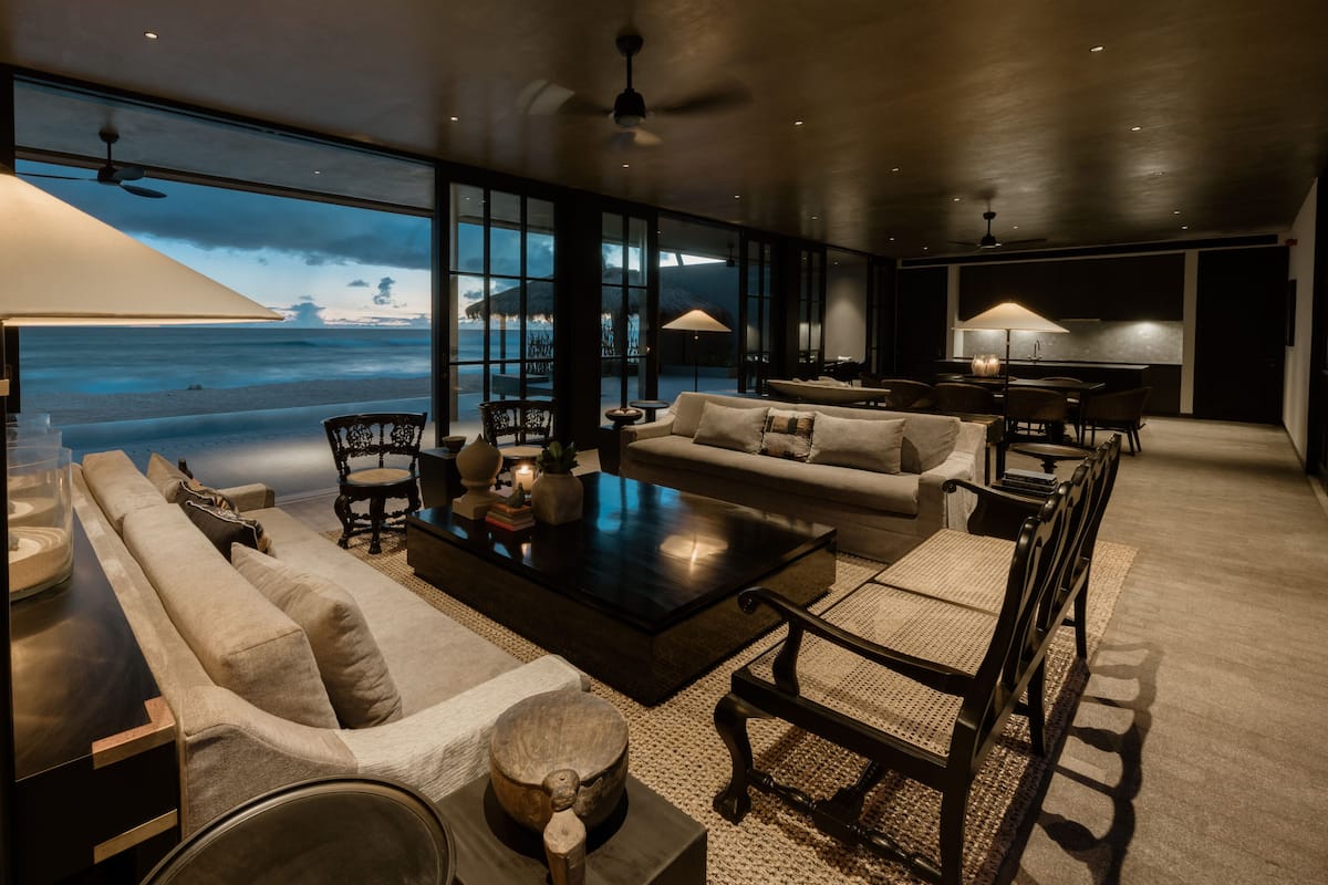 A modern living room with a large window overlooking a beach, furnished with sofas, a coffee table, and lamps.