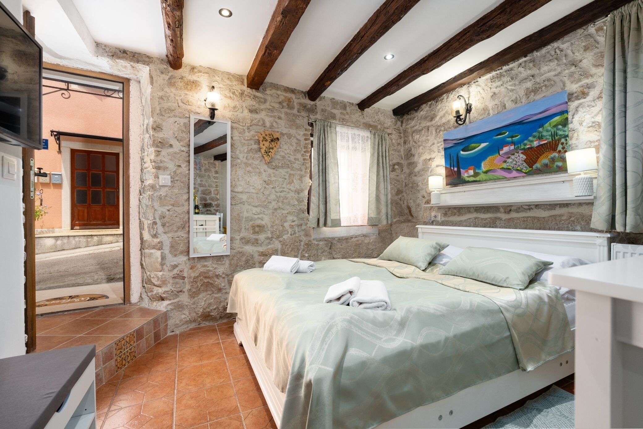 Un dormitorio con pared de piedra, una cama con colcha verde, un cuadro sobre la cama y una ventana con cortinas.
