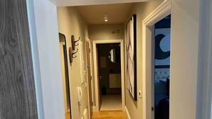 A hallway with wooden flooring, framed artwork on the walls, and a door leading to a bedroom with a tufted headboard. - Stunning apartment by The Leas, West End (Folkestone)