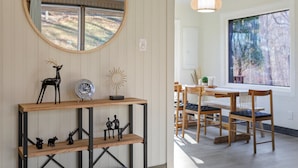 A modern interior with a wooden console table, a round mirror, and a dining area with wooden chairs and a table.
