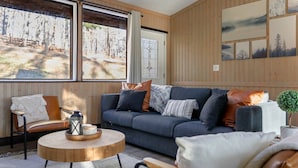 A cozy living room with a blue sofa, wooden walls, and large windows offering a view of a forest.