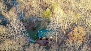 An aerial view of a cabin surrounded by trees, with a deck and a covered porch.