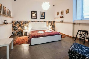 Un dormitorio moderno con una cama grande, cabecera blanca y un panel decorativo en la pared. Hay mesitas de noche con lámparas, un pequeño taburete y una ventana con vistas.