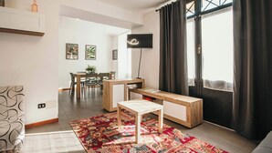 Ein modernes Wohnzimmer mit einer Couch, einem Couchtisch, einem Fernseher, einem Essbereich mit Stühlen und einem Tisch sowie einem großen Fenster mit Vorhängen.
