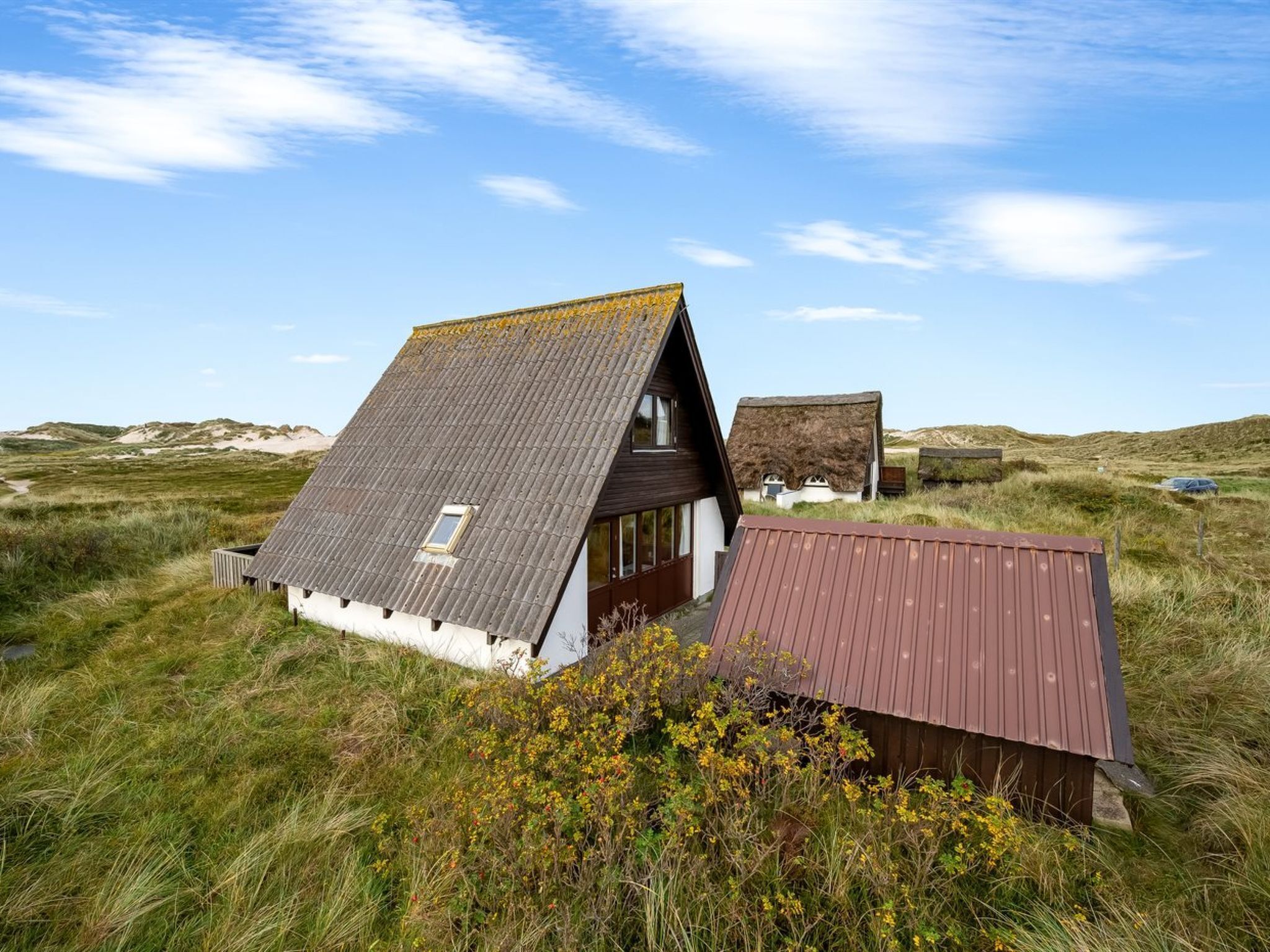 A traditional house with a sloped roof, surrounded by grassy dunes and a clear sky.