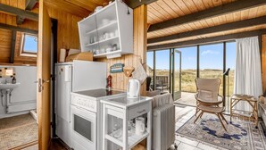 A compact kitchen with a refrigerator, stove, and open shelving. A dining area with a chair and table is visible through a glass door, overlooking a natural landscape. - "Fausta" - 150m from the sea by Interhome (Ringkøbing)