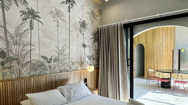 Hotelkamer met een groot bed, een houten hoofdbord, een tropische muurschildering, een moderne lamp en uitzicht op het buitenterrein met zitplaatsen.