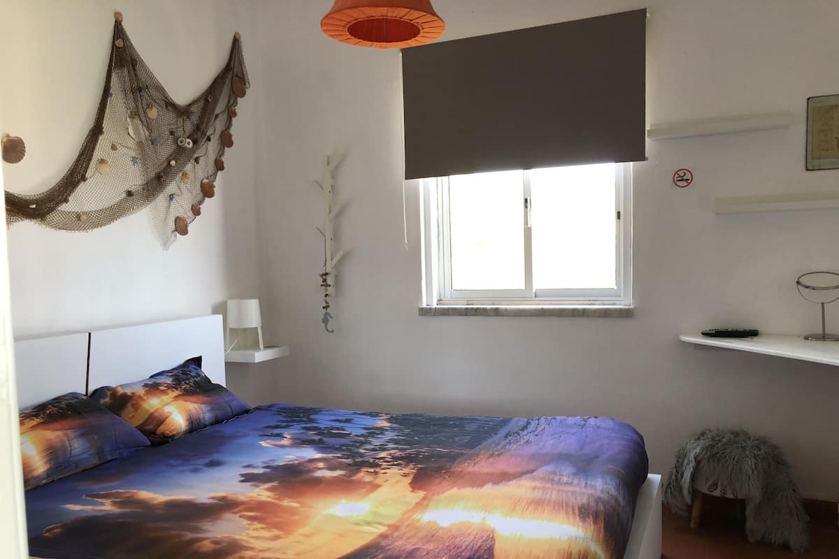 A bedroom with a bed, a window, a hanging decoration, and a small shelf.