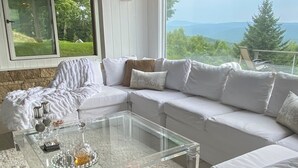 A modern living room with a glass coffee table, white sofas, and a view of a mountainous landscape through large windows.