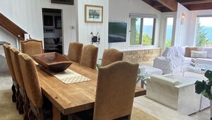 A spacious living room with a wooden dining table, comfortable seating, a stone fireplace, and a large window offering a view of the outdoors.