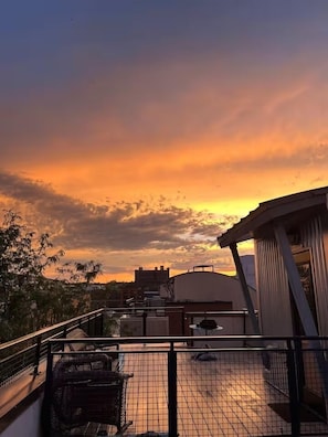 Terraço com vista para o pôr do sol, com guarda-corpo de metal e um pequeno espaço para sentar.
