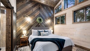 A bedroom with a hipped roof, a large bed, a nightstand, and a window with a view of trees.