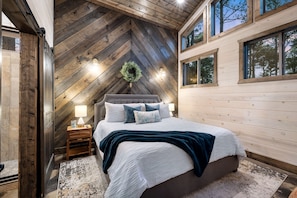 A bedroom with a hipped roof, a large bed, a nightstand, and a window with a view of trees.