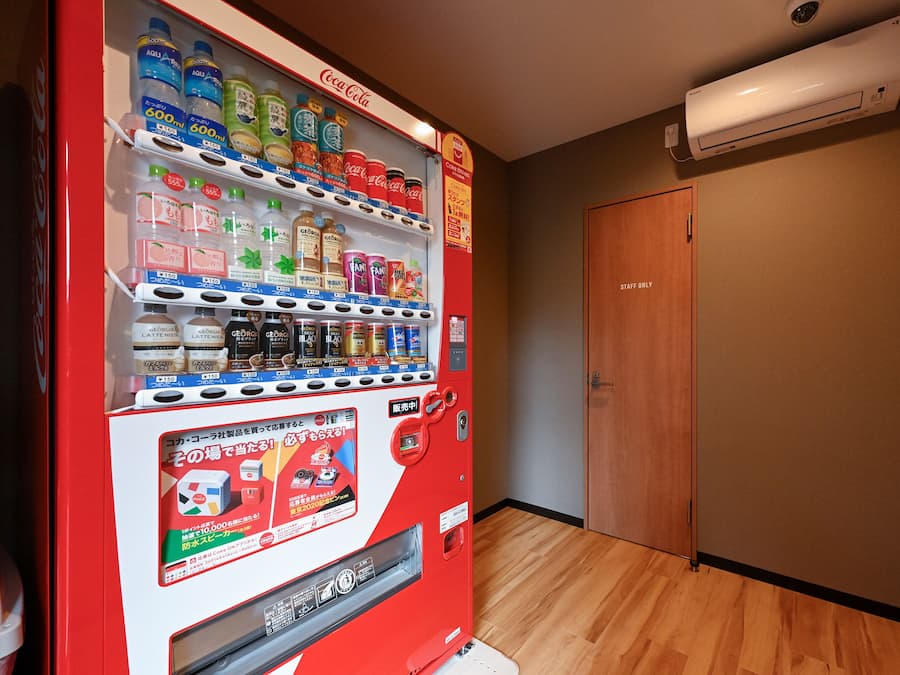 A vending machine stocked with beverages and snacks, a wooden door, and an air conditioning unit on the wall.