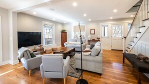 A spacious living room with a large flat-screen TV, grey sofas, a coffee table, and a staircase leading to another level. - Sunsets On Blue Hill, Newly Renovated Farmhouse (Great Barrington)