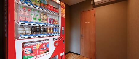 A vending machine stocked with beverages and snacks, a wooden door, and an air conditioning unit on the wall.