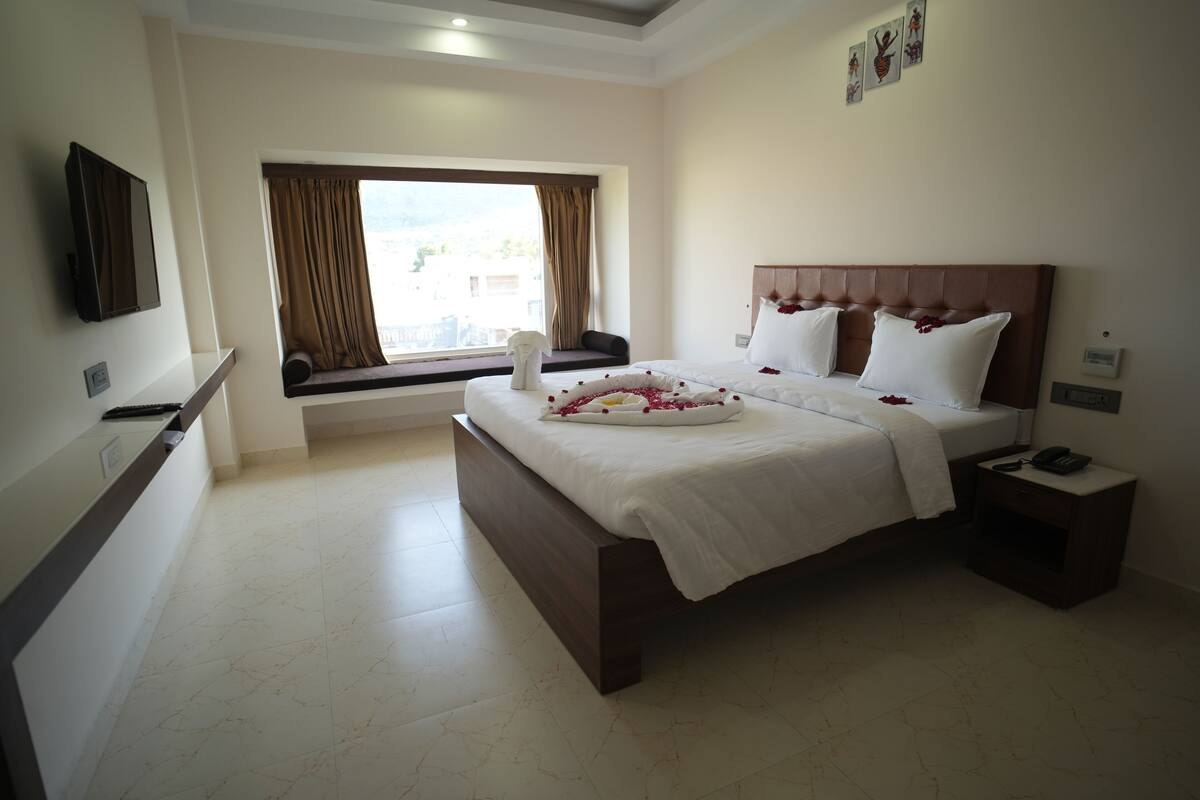 Quarto de hotel com uma cama grande, uma mesinha de cabeceira, uma TV fixada na parede e uma janela com cortinas.