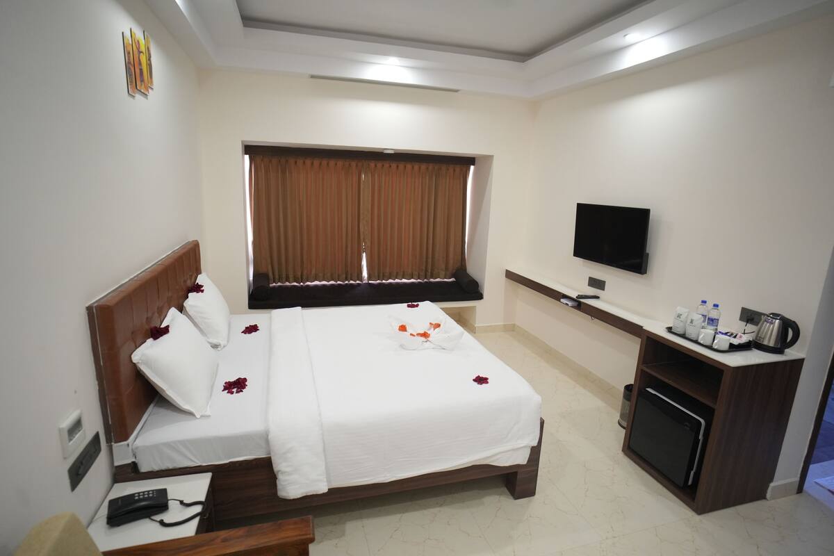 Quarto de hotel com cama, televisão, mesa de trabalho com máquina de café e janela com cortinas.
