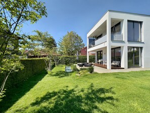 Une maison moderne à deux étages, dotée d’une pelouse bien entretenue, d’un espace extérieur aménagé pour se détendre et d’un grand balcon vitré.