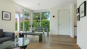En moderne stue med et glasbord, en sofa, en bænk og et spiseområde med en bænk. Der er en dør, der fører til en balkon med udsigt over grønne områder.