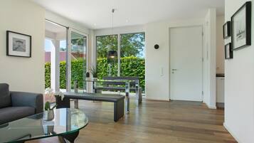 Ein modernes Wohnzimmer mit einem Glastisch, einer Couch, einer Bank und einem Essbereich mit einer Bank. Eine Tür führt zu einem Balkon mit Blick ins Grüne.