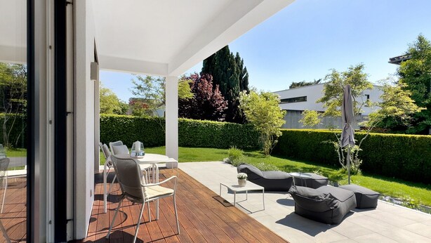 En udendørs patio med træterrasse, hvide vægge og udsigt til en velholdt have og moderne bygninger.