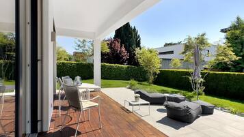Eine Terrasse im Freien mit Holzdeck, weißen Wänden und Blick auf einen gepflegten Garten sowie moderne Gebäude.