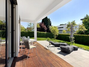 Une terrasse extérieure avec une plateforme en bois, des murs blancs et une vue sur un jardin bien entretenu et des bâtiments modernes.