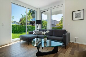 Un salon moderne avec une table basse en verre, un canapé gris et une vue sur un jardin grâce à de grandes fenêtres.