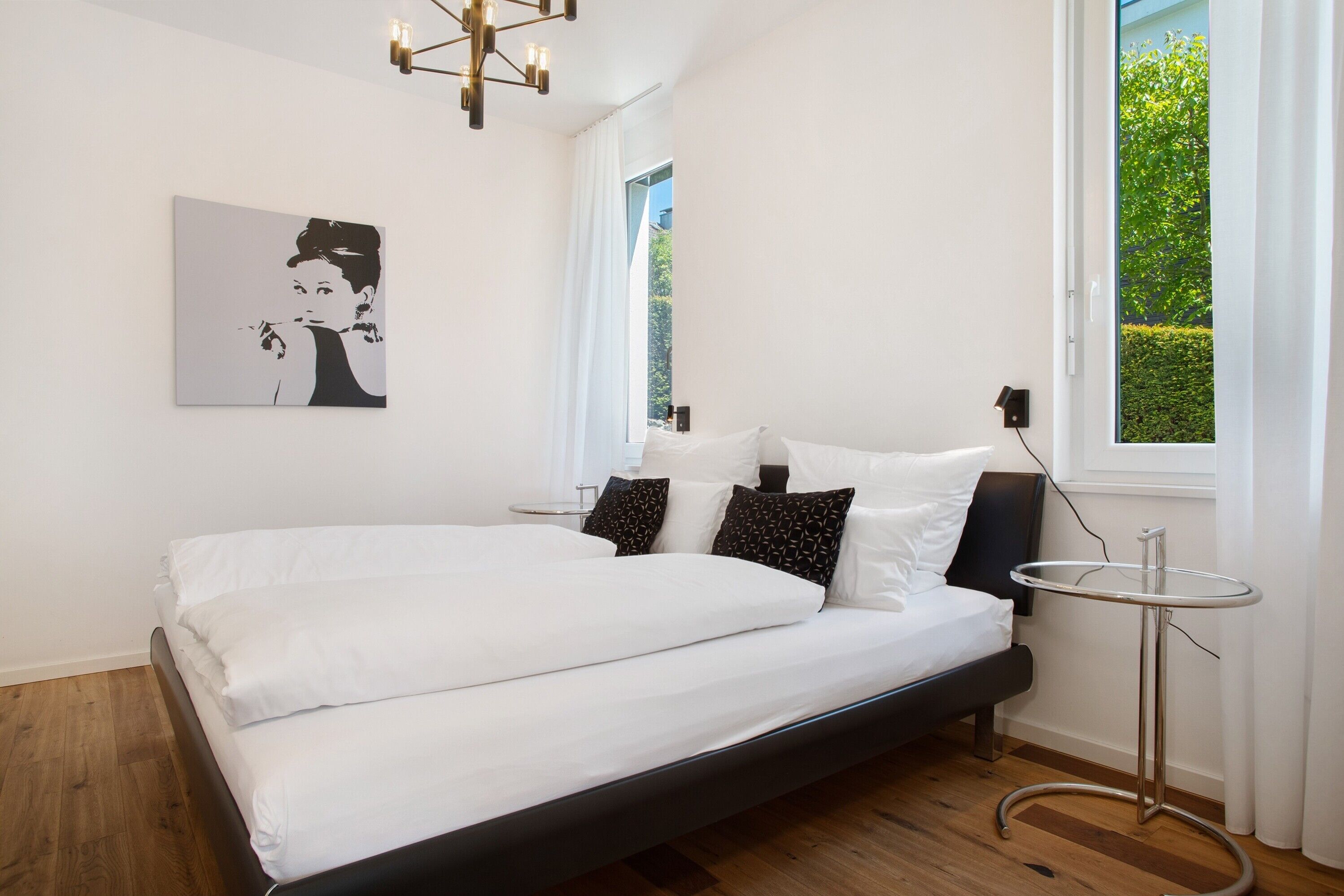 Ein modernes Schlafzimmer mit Bett, Nachttisch, Wandleuchte, einem Fenster mit Vorhängen und einem gerahmten Bild an der Wand.