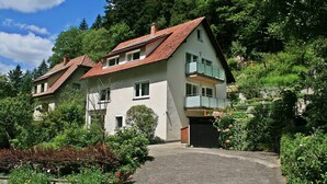Une maison à deux étages avec un toit en pente, un balcon à l’étage supérieur et une allée menant à la porte d’entrée.
