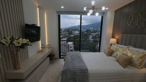 Un dormitorio moderno con una cama grande, un televisor montado en la pared, un banco con objetos decorativos y un balcón con vista a la ciudad.