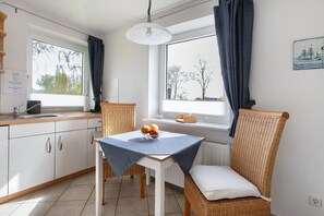 Uma cozinha com área de refeições, uma janela com cortinas, uma cadeira de vime e uma mesa com uma tigela de frutas.