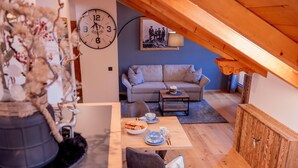 Een knus loft-achtig vertrek met een houten plafond, een bank, een eettafel met een schaal koekjes en een klok aan de muur.