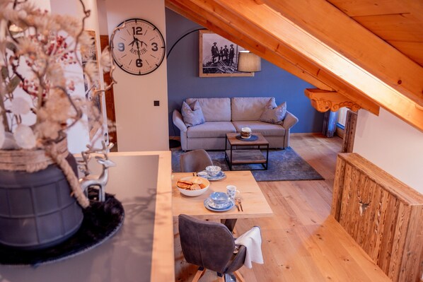 A cozy loft-style room with a wooden ceiling, a sofa, a dining table with a bowl of cookies, and a clock on the wall.
