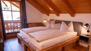 Een bed met houten frame, beddengoed met ruitpatroon, een nachtkastje met lamp en een raam met gordijnen.