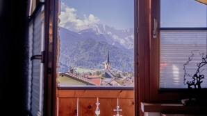 Een kamer met houten inrichting, een raam met uitzicht op een berg en een kerk, en een klein tafeltje met een vaas.