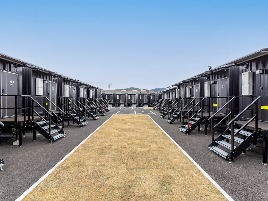 Eine Reihe identischer Hotelzimmer mit Nummern auf den Türen, einem zentralen Gang und blauem Himmel.