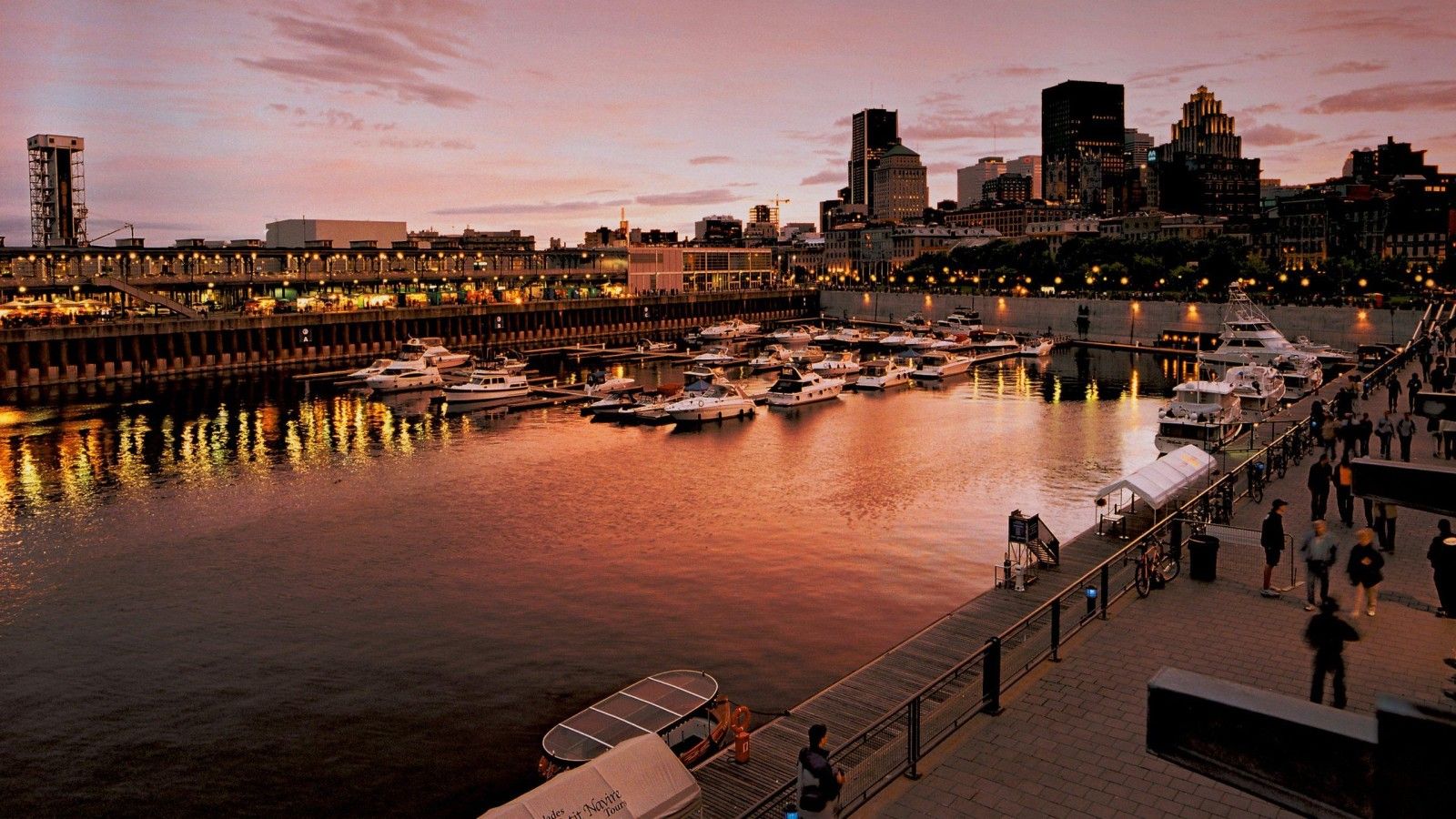 A waterfront marina with boats docked, a city skyline in the background, and a pedestrian walkway along the water.