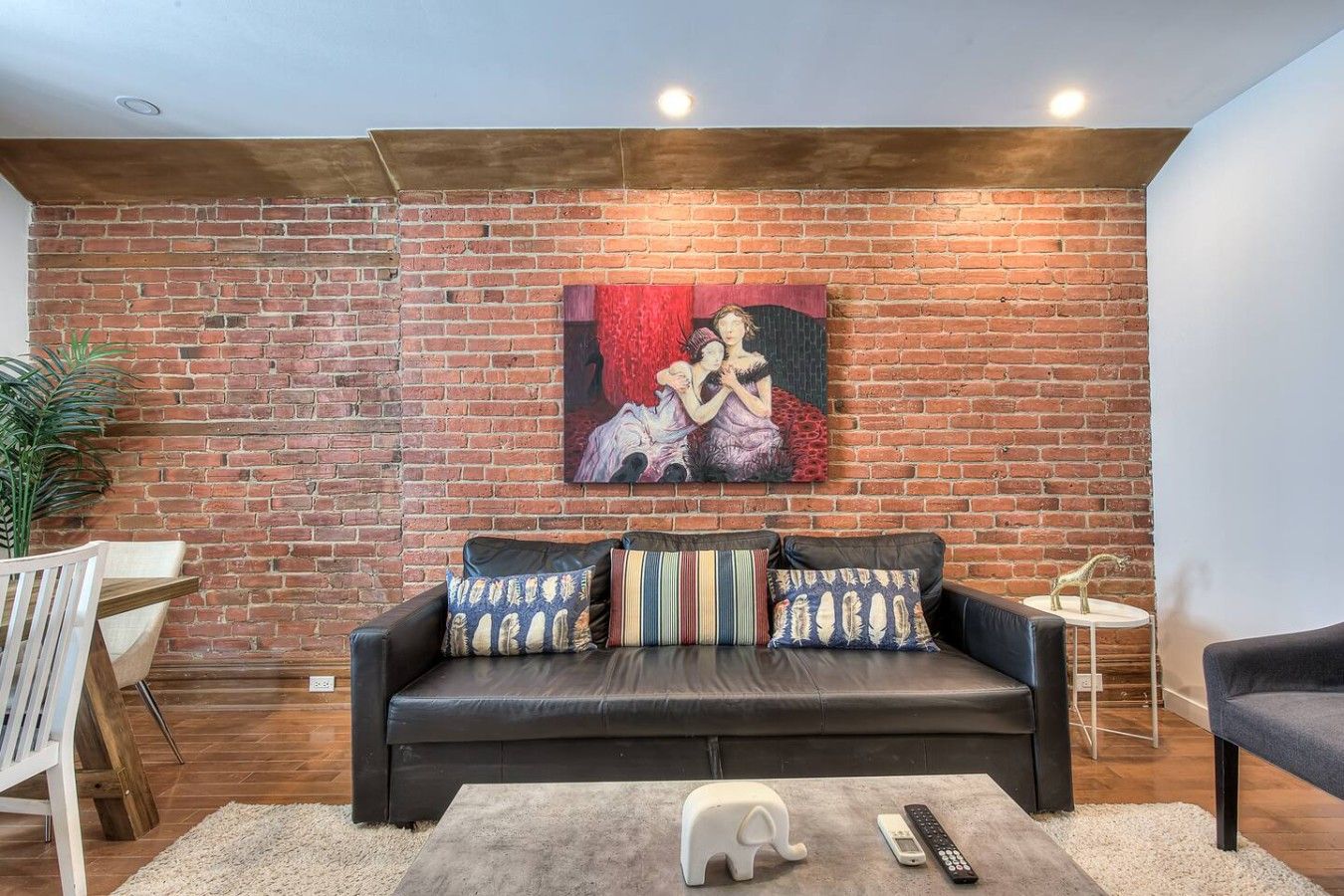 A modern living room with a brick wall, a black leather sofa, colorful cushions, a white elephant-shaped lamp, and a painting above the sofa.