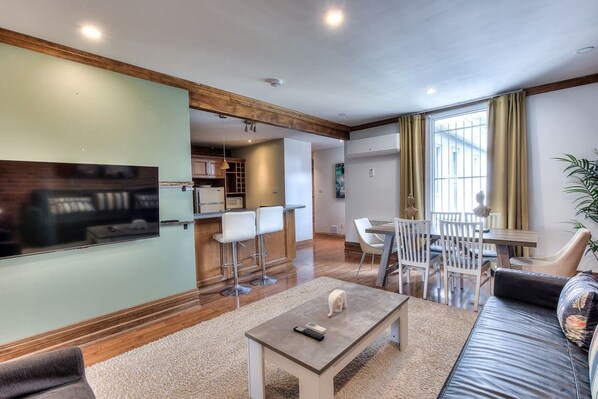 Una sala de estar moderna con un televisor de pantalla plana, una zona de comedor con sillas y una cocina al fondo.