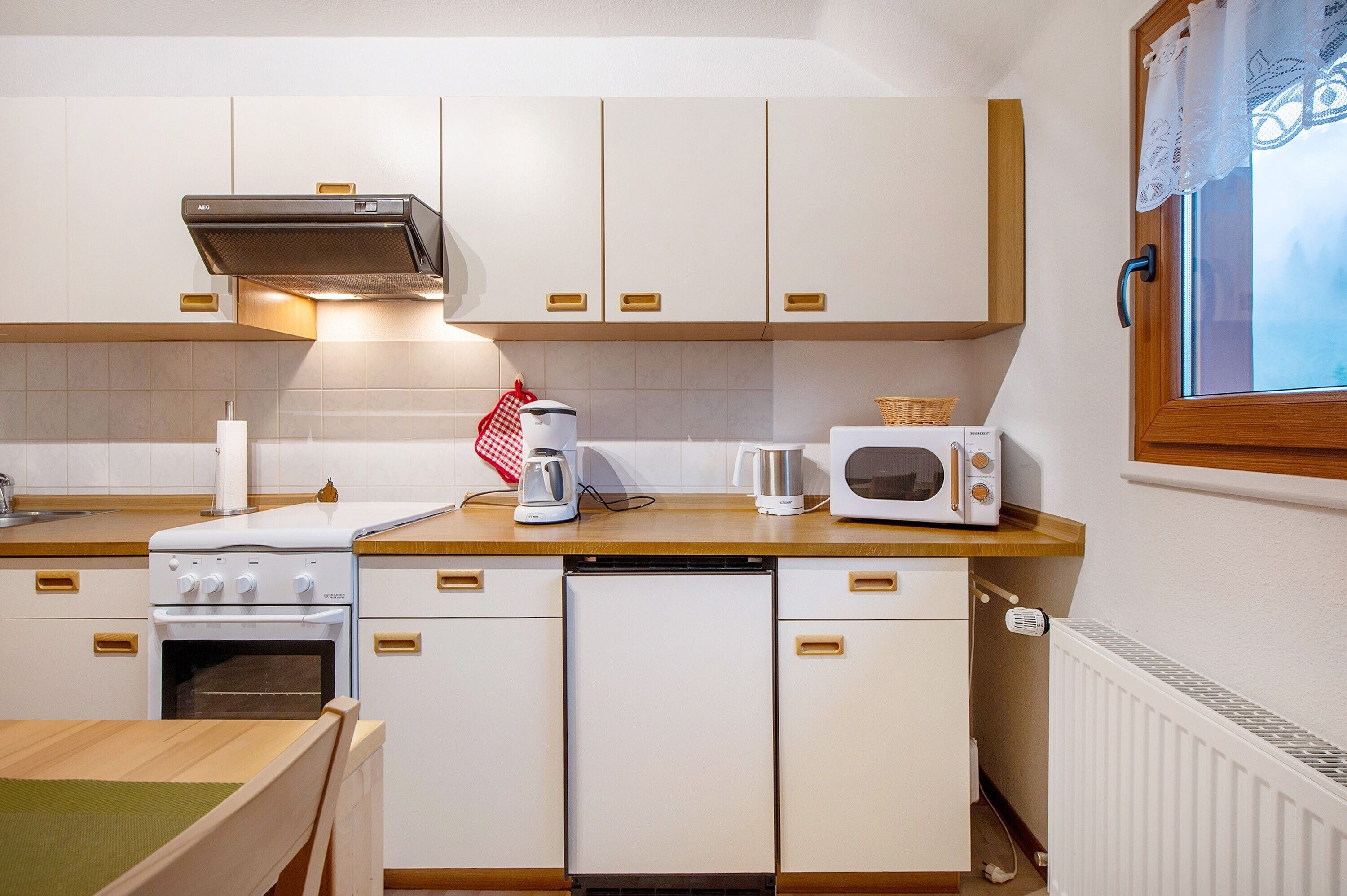 Une cuisine compacte avec des meubles blancs, une cuisinière, un lave-vaisselle, un four à micro-ondes et une fenêtre ornée de rideaux en dentelle.