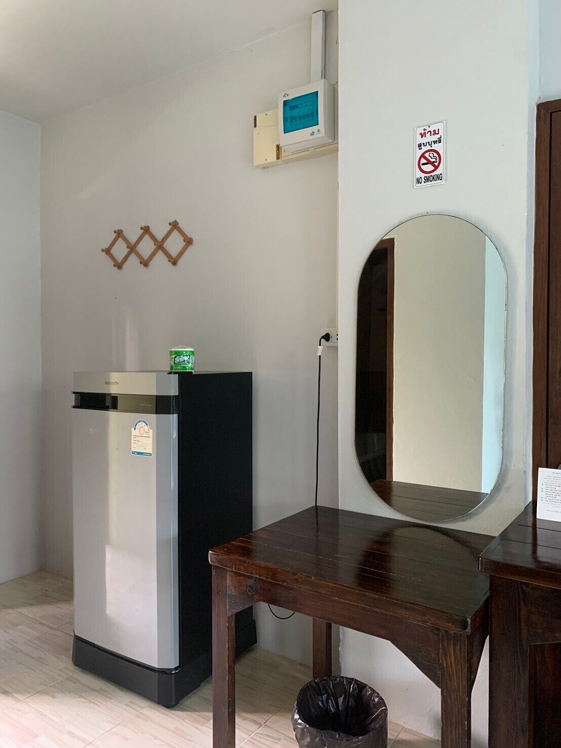 A compact room with a refrigerator, a small table, a trash bin, a wall-mounted TV, and a no-smoking sign.
