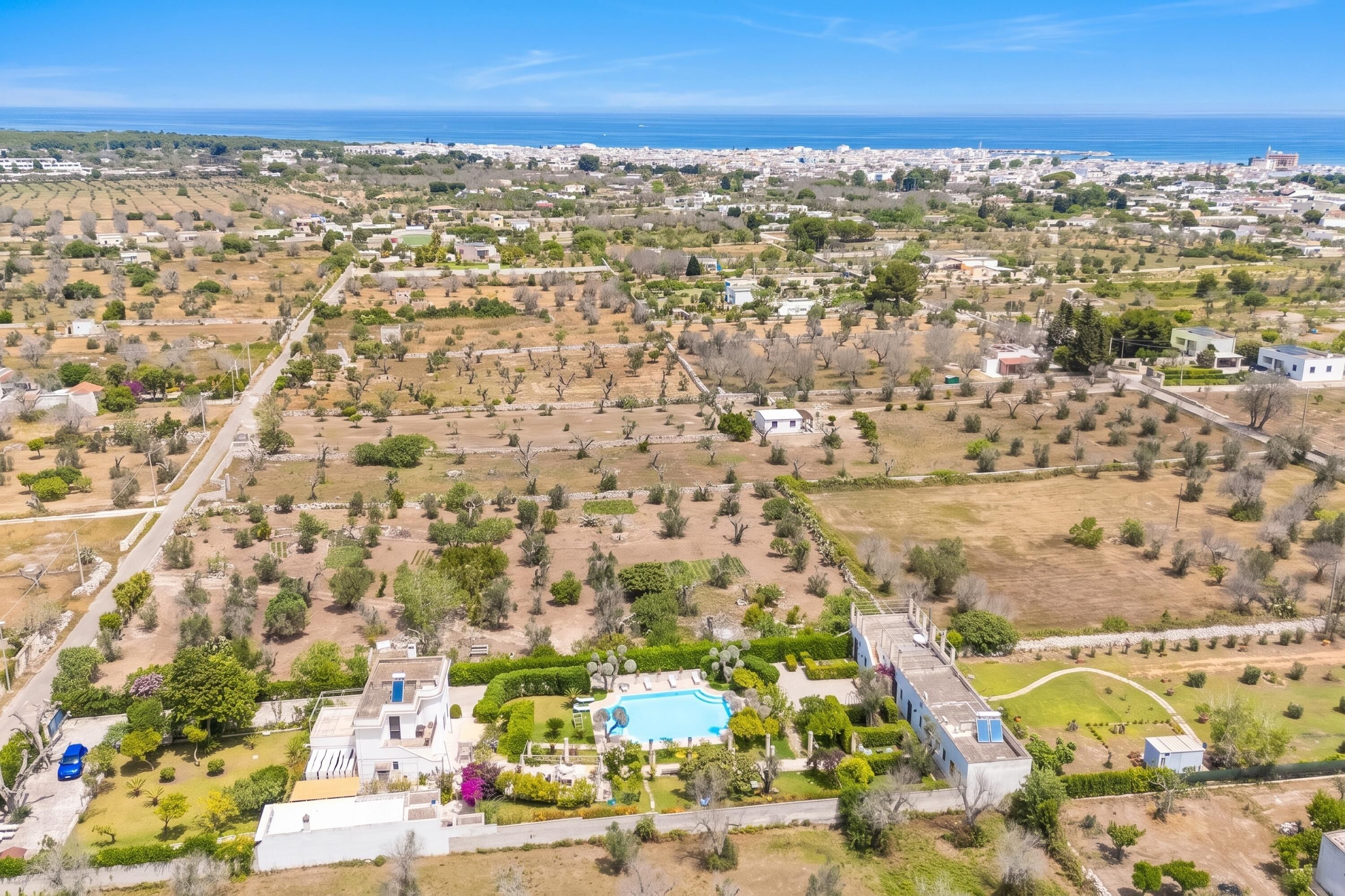 An aerial view of a residential area with a large swimming pool surrounded by greenery, a clear blue sky, and a distant view of the sea.