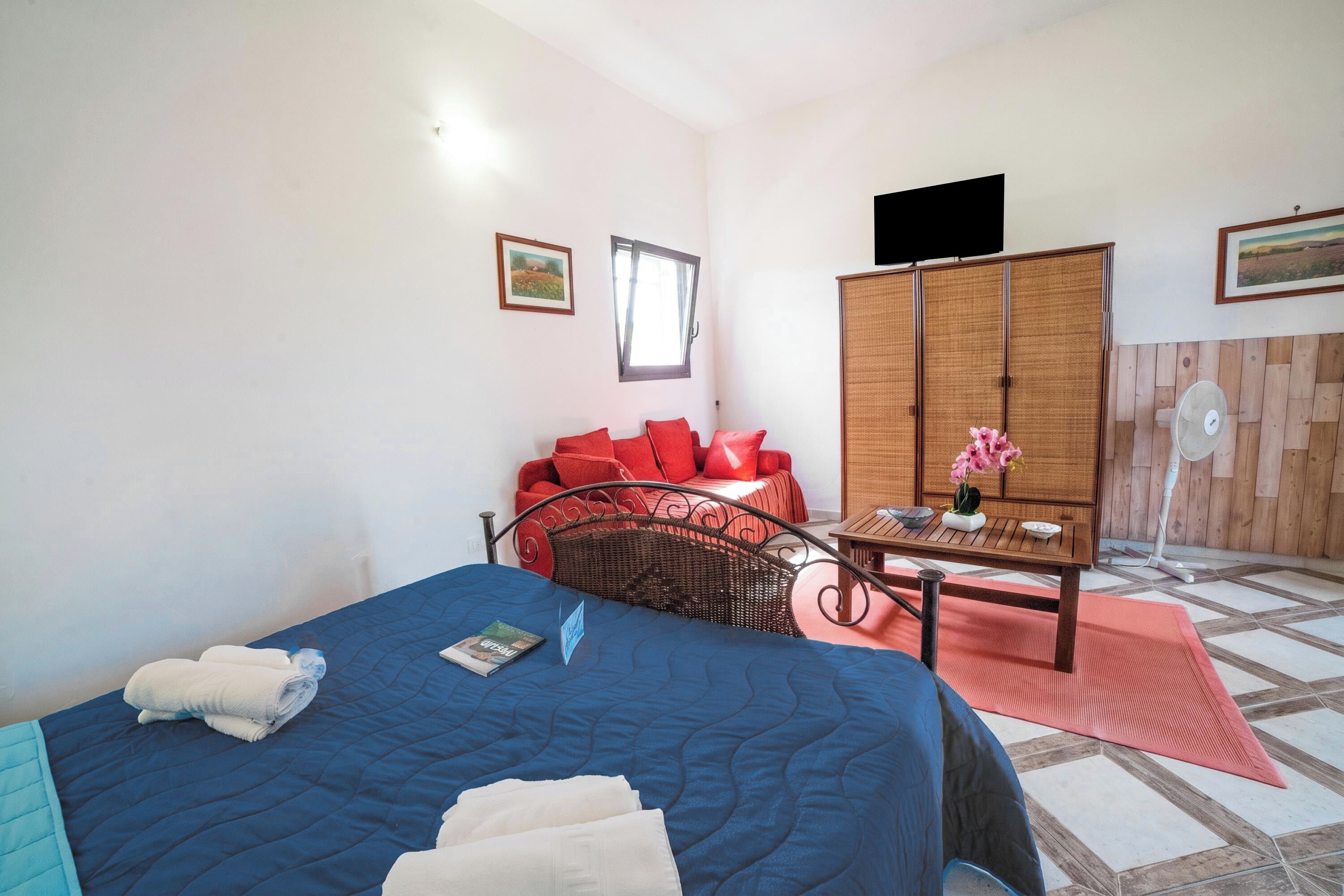 A bedroom with a bed, a red sofa, a wooden wardrobe, a small table with a vase, and a fan.