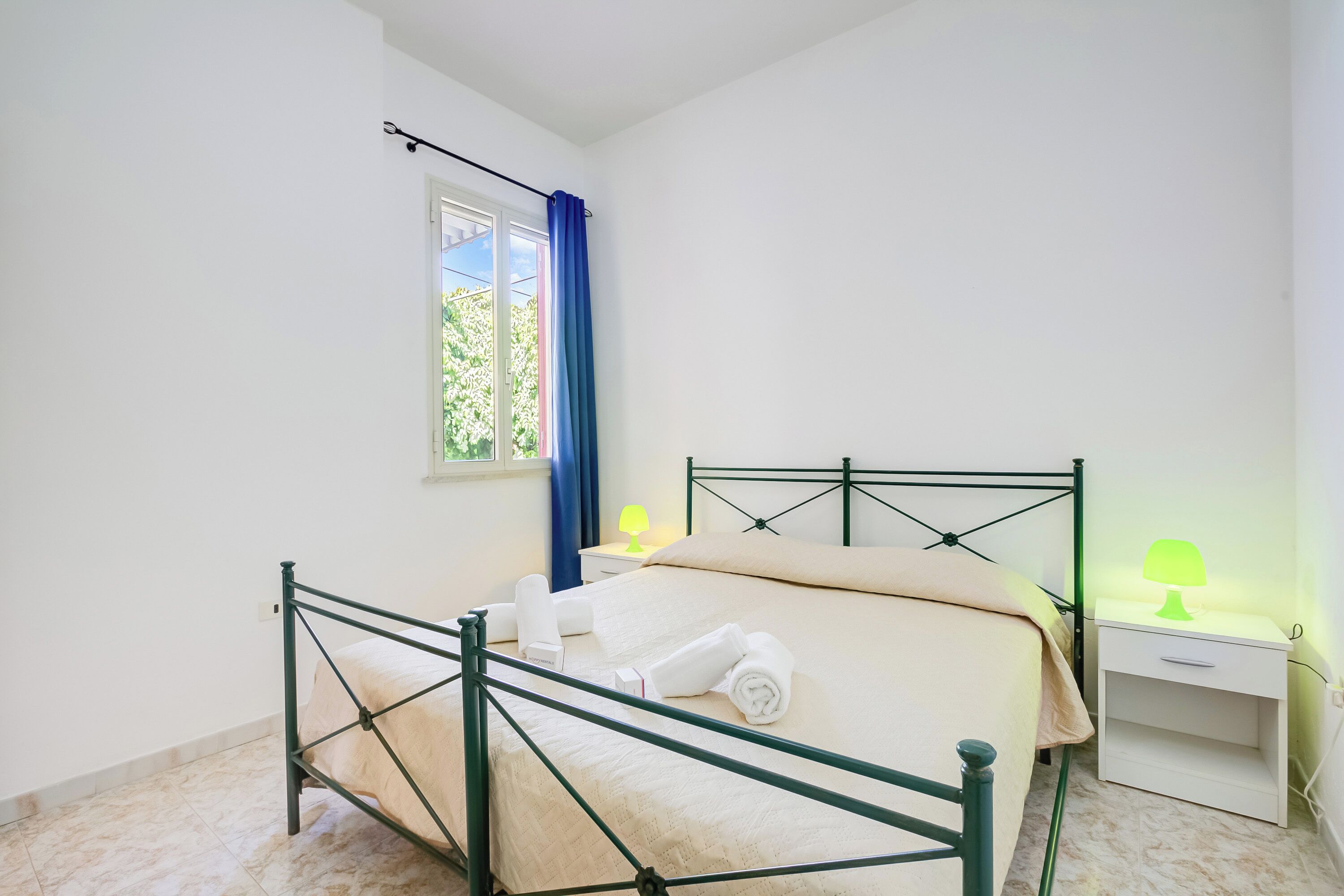 A bedroom with a bed, bedside tables, a window with curtains, and a view of greenery outside.